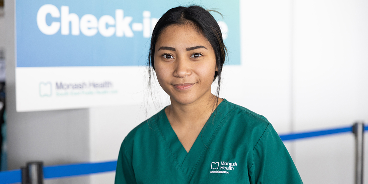 Asiah Vergara stands in front of a blue Check-in sign. She's smiling and wearing forest green scrubs with her black hair pulled back into a ponytail.