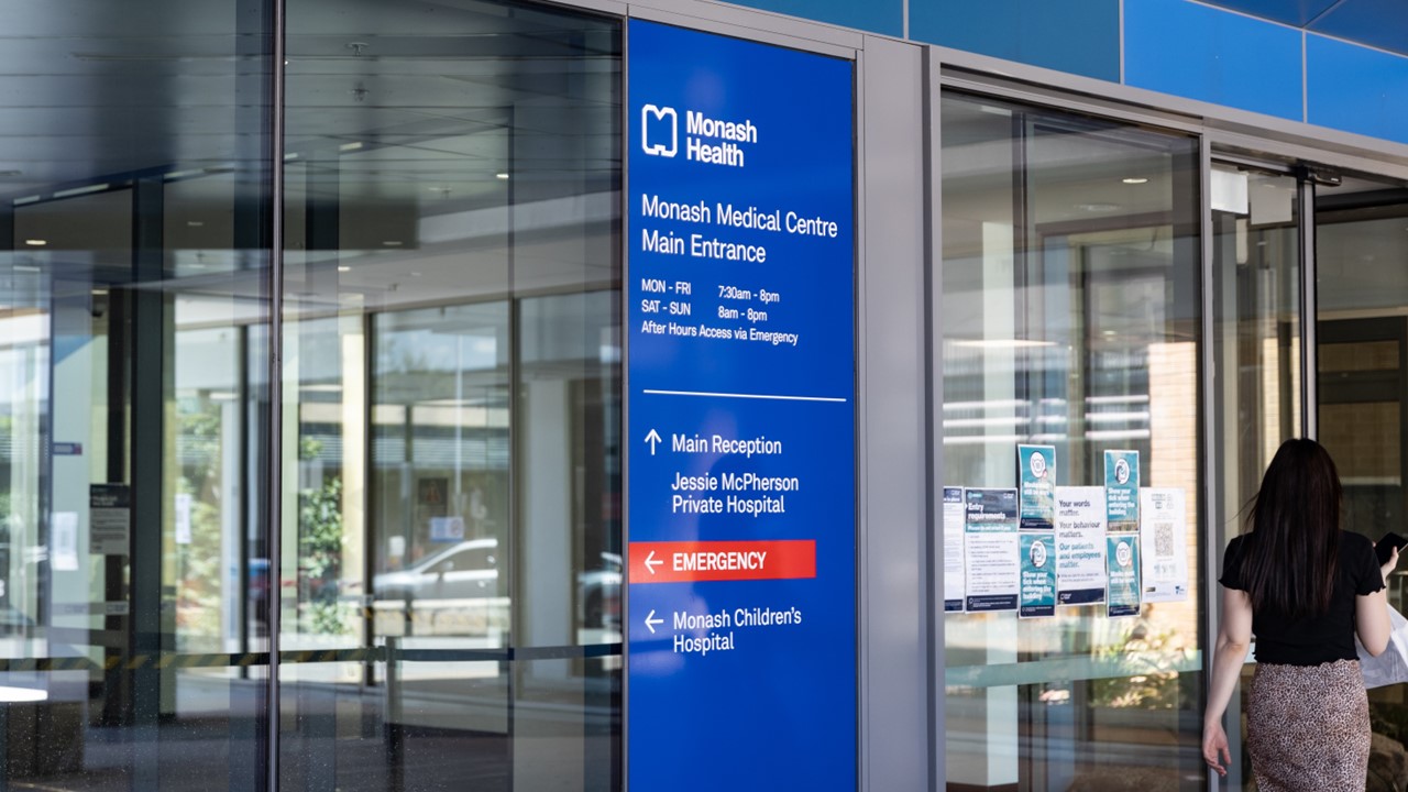 Monash Health branded signage at the entrance of the hospital, and a woman walking in through the glass doors.