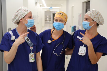 Photo of two clinical employees pointing to Dr Jackson between them, while wearing their colourful cloth hats in the ward