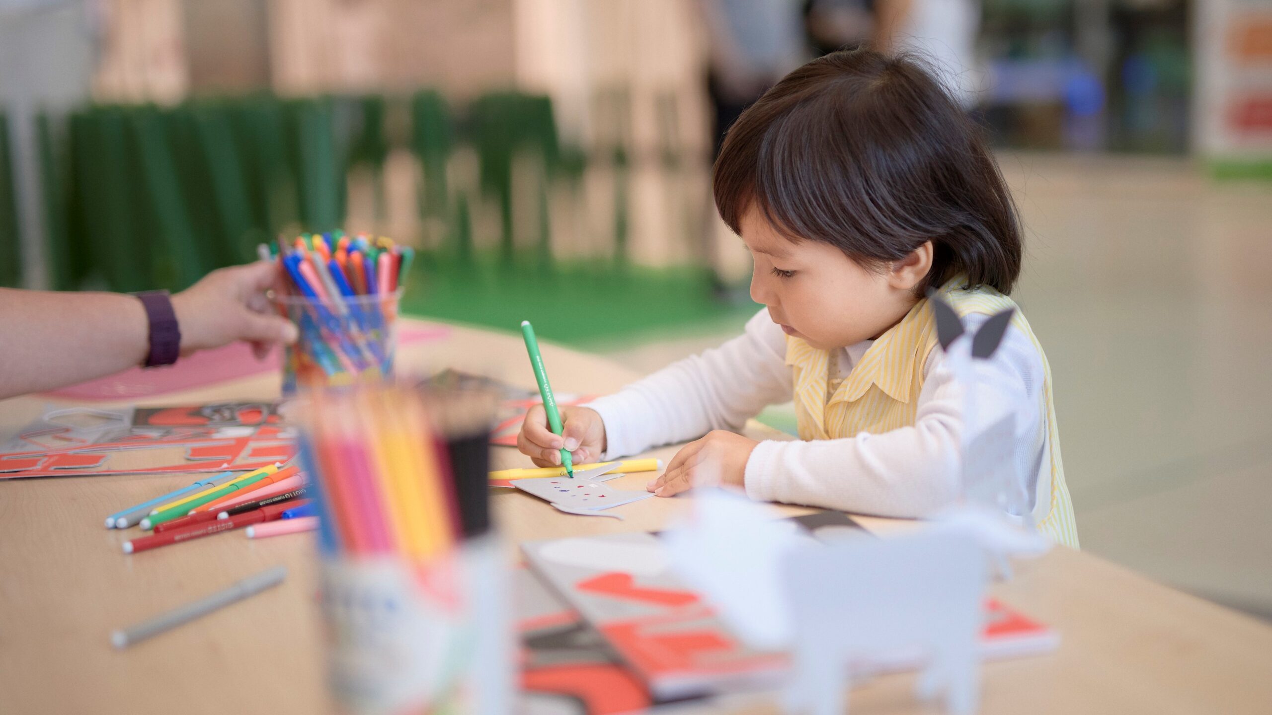 Young child with dark hair drawing, surrounded by colourful pencils and markers