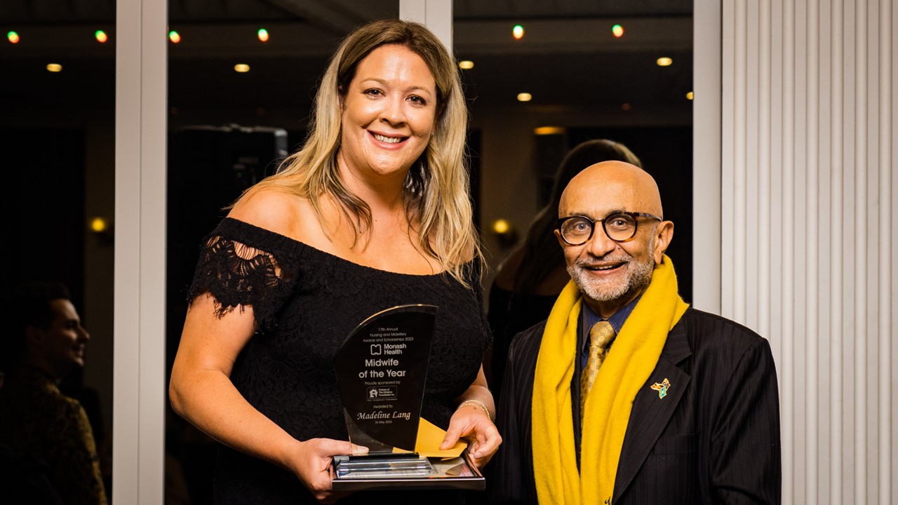 Madeline Lang stands next to Board Chair Dipak Sanghvi. She wears an off the shoulder black dress, has mid-length blonde hair and is holding her Midwife of the Year photo,