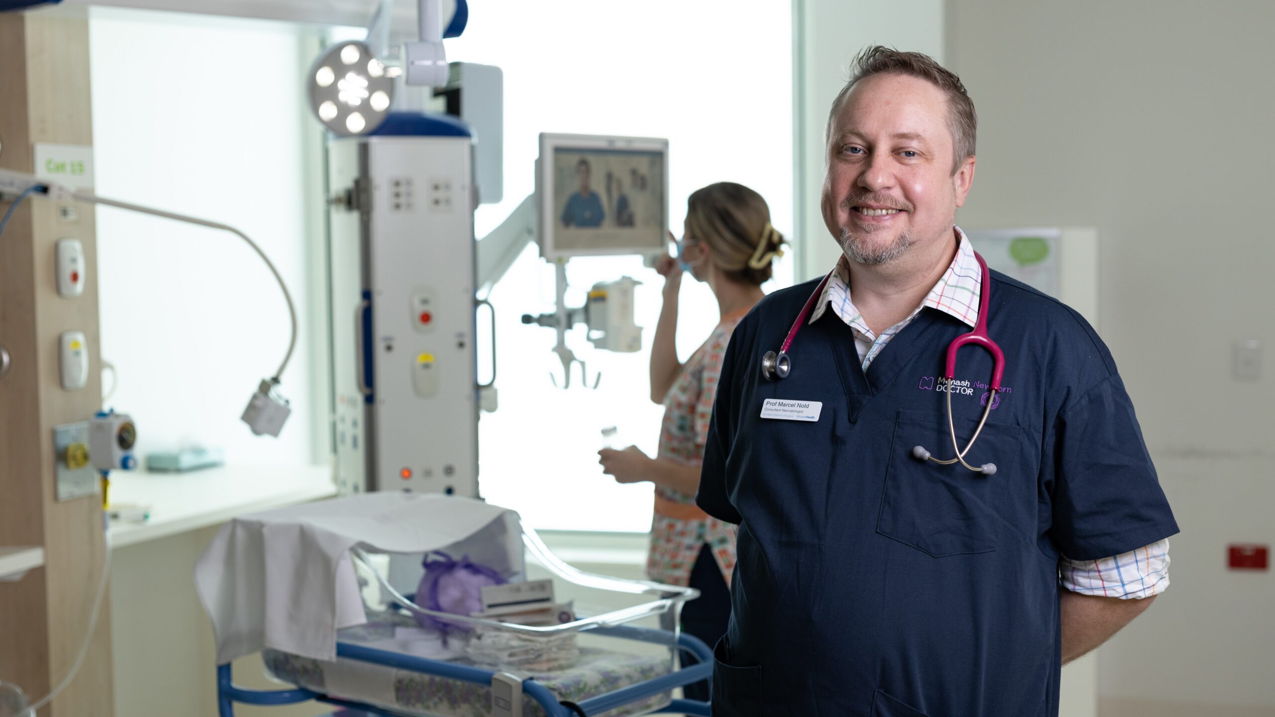 Professor Marcel Nold smiles in front of a baby cot and a nurse looking at a monitor.