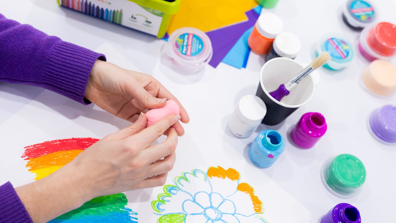 A pair of hands plays with some putty on a table covered in art equipment including paints and pencils.