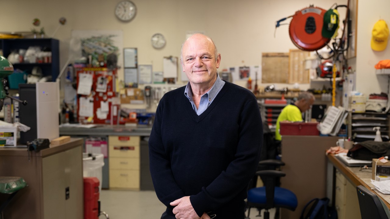 A man wearing a blue shirt and dark blue jumper standing in a workshop, smiling to camera
