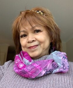 A lady with short auburn hair wearing a lavendar top with a purple scarf, smiling to camera