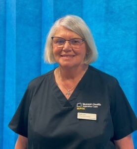 A lady with short grey hair wearing nurse scrubs stands in front of a blue divider curtain
