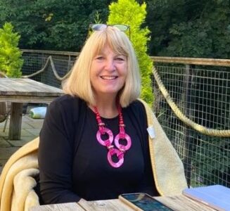 A lady with short blonde hair wearing a black top with a large pink necklace sitting at an outdoor table, smiling to camera