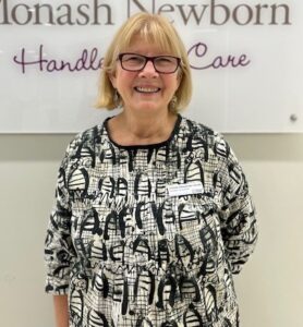 A lady with short blonde hair wearing a black and white top standing in front of the Monash Newborn sign, smiling to camera