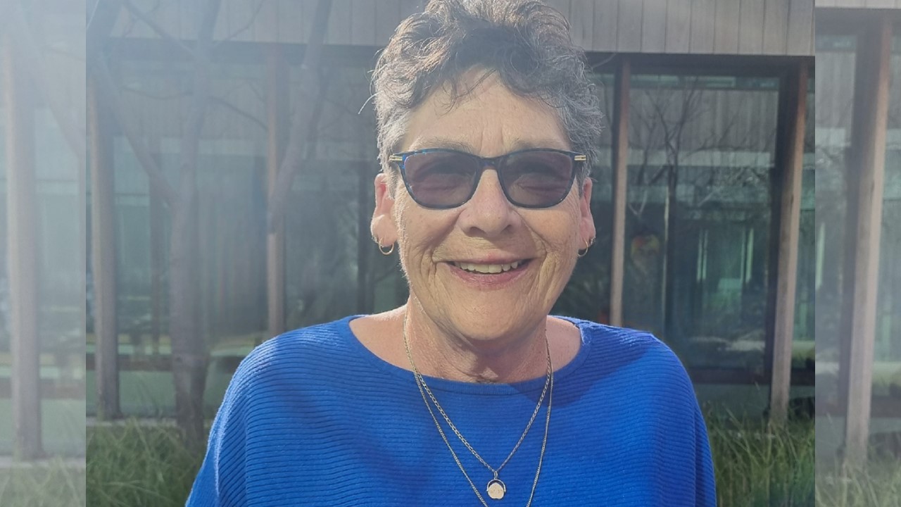 A lady wearing sunglasses and a blue top smiles to camera