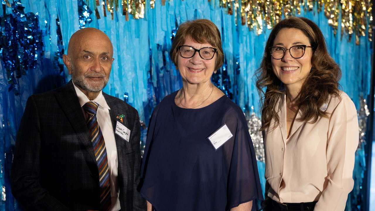 A man and two ladies standing side-by-side, smiling to camera in front of a tinsel backdrop