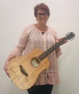 A lady wearing a light pink top holds an acoustic guitar and smiles to camera