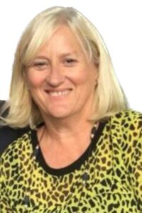 A lady with shoulder-length blonde hair wearing a yellow leopard-print top smiles to camera