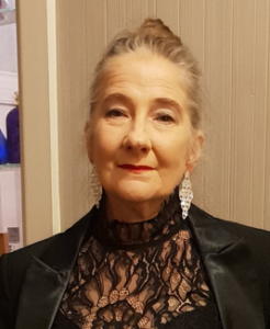 A lady with light-coloured hair pulled back, wearing a black lacy top and silver dangly earrings, looking at the camera