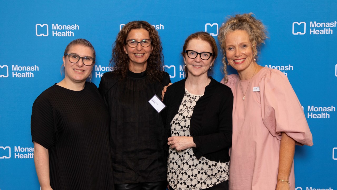 Four ladies smile to camera, standing in front of a blue Monash Health media wall