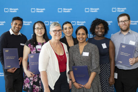 Seven people smile to camera, standing in front of a blue Monash Health media wall