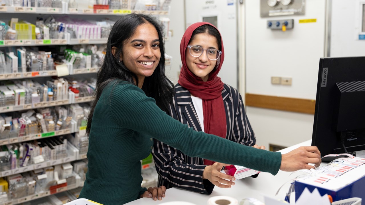 Two young ladies standing at a computing looking to camera, against a backdrop of shelves of pharmaceuticals