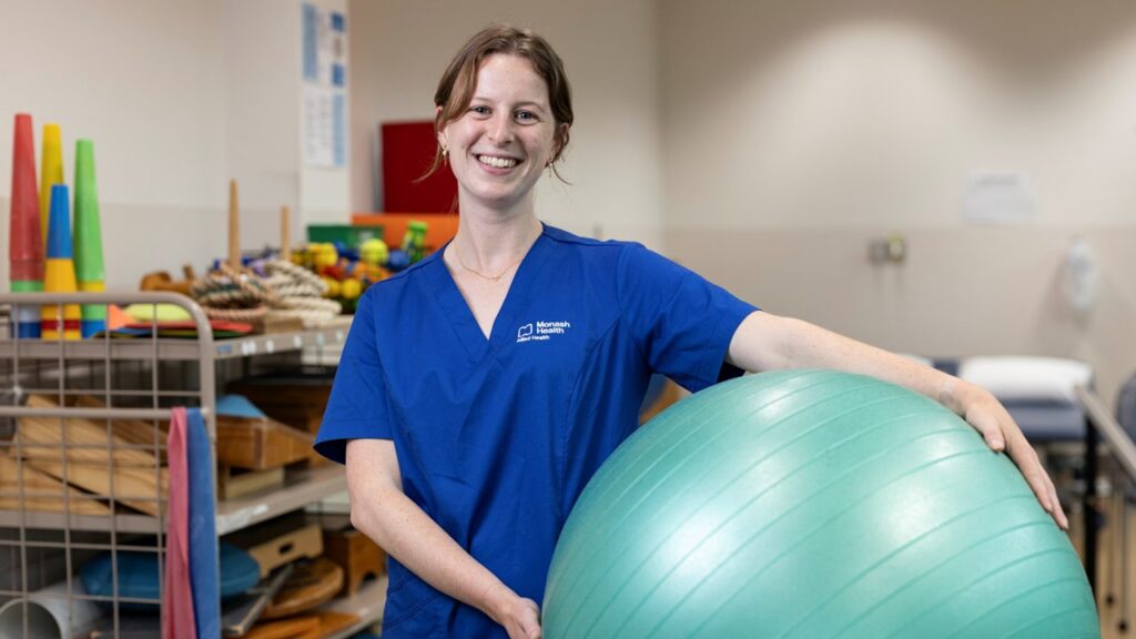 Sophie Clarke 1 1920x1080px Sophie Clarke stands in a gym holding a large green yoga ball while smiling.
