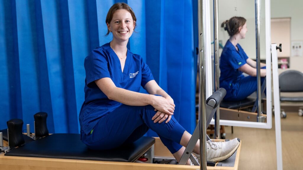 Sophie Clarke 3 1920x1080px Sophie sits on a reformer pilates machine and is smiling.